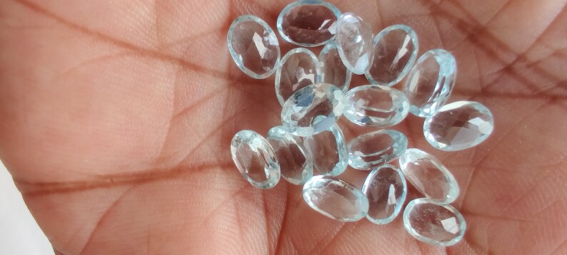 May include: A scattering of oval-cut, pale blue gemstones. The faceted stones are displayed on a palm, highlighting their clarity and colour. These gemstones are likely for jewellery making.