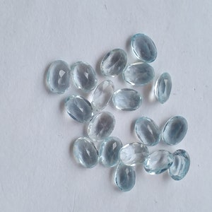 May include: A scattering of oval, pale blue gemstones on a white background. The translucent stones have a subtle shimmer. The arrangement is random, giving a natural appearance. The gemstones are likely aquamarine.