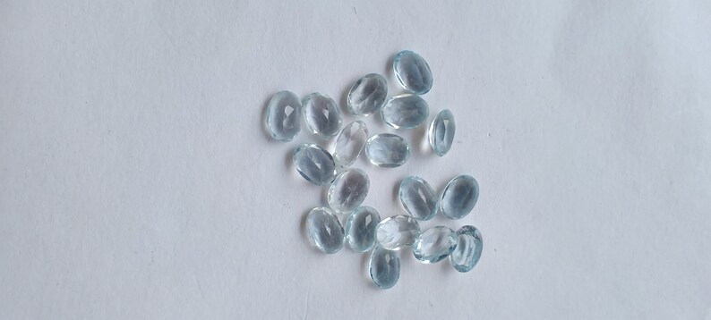 May include: A scattering of oval, pale blue gemstones on a white background. The translucent stones have a subtle shimmer. The arrangement is random, giving a natural appearance. The gemstones are likely aquamarine.