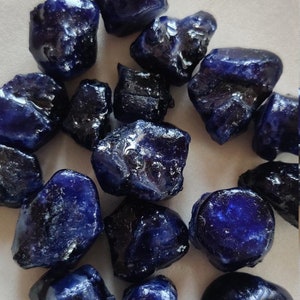 Puede incluir: Una colección de piedras preciosas de zafiro azul oscuro en bruto. Las piedras preciosas son de varios tamaños y formas, algunas tienen un ligero brillo iridiscente.