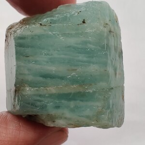 Pode incluir: Um cristal de &aacute;gua-marinha azul claro com uma textura &aacute;spera e natural. O cristal tem uma forma retangular e &eacute; transl&uacute;cido.