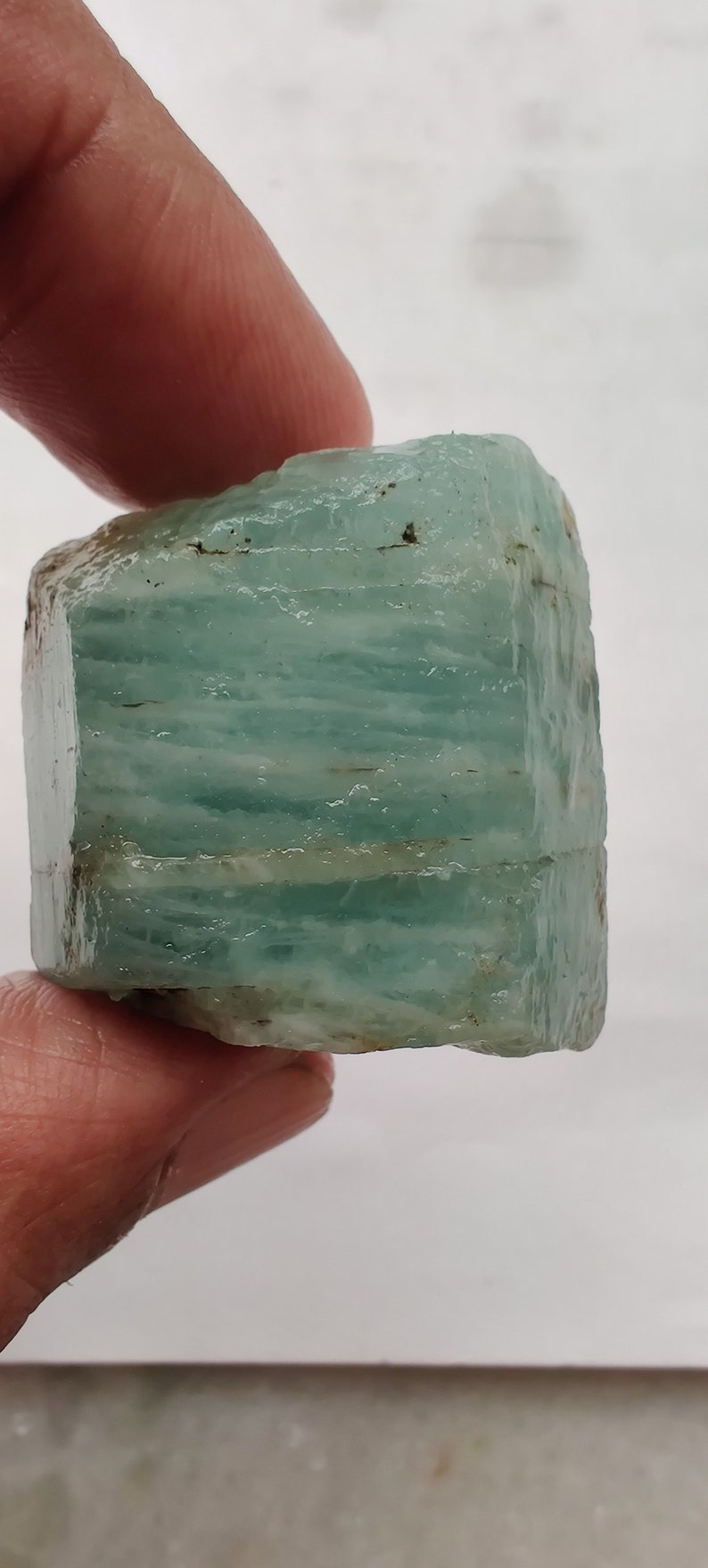 Pode incluir: Um cristal de &aacute;gua-marinha azul claro com uma textura &aacute;spera e natural. O cristal tem uma forma retangular e &eacute; transl&uacute;cido.