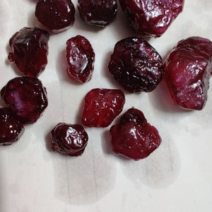 May include: A collection of 12 deep red ruby gemstones of various sizes and shapes. The gemstones are scattered on a white surface.
