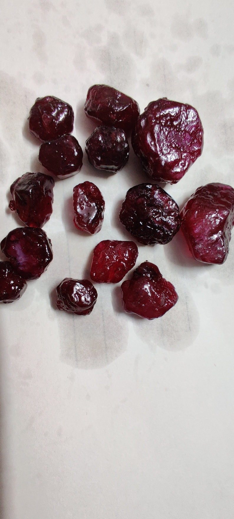 May include: A collection of 12 deep red ruby gemstones of various sizes and shapes. The gemstones are scattered on a white surface.
