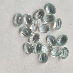 May include: A scattering of oval, pale blue gemstones on a white background. The gemstones are translucent and faceted, reflecting light. The image showcases a collection of gemstones, suitable for jewellery.