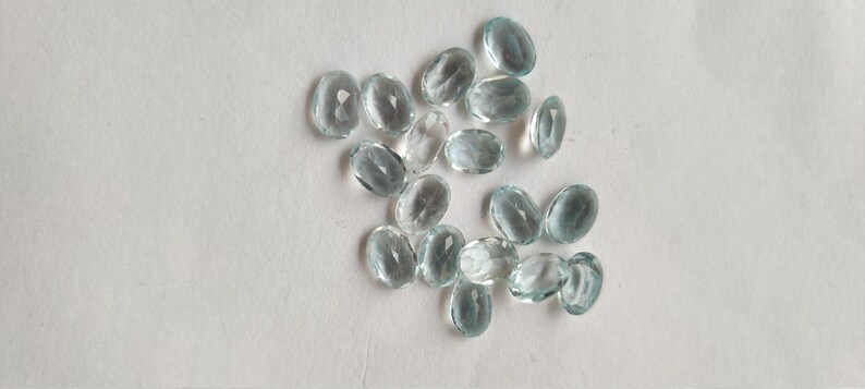 May include: A scattering of oval, pale blue gemstones on a white background. The gemstones are translucent and faceted, reflecting light. The image showcases a collection of gemstones, suitable for jewellery.