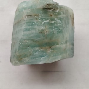 Pode incluir: Um cristal de &aacute;gua-marinha azul claro com uma textura &aacute;spera e natural. O cristal tem uma apar&ecirc;ncia em camadas com estrias vis&iacute;veis.