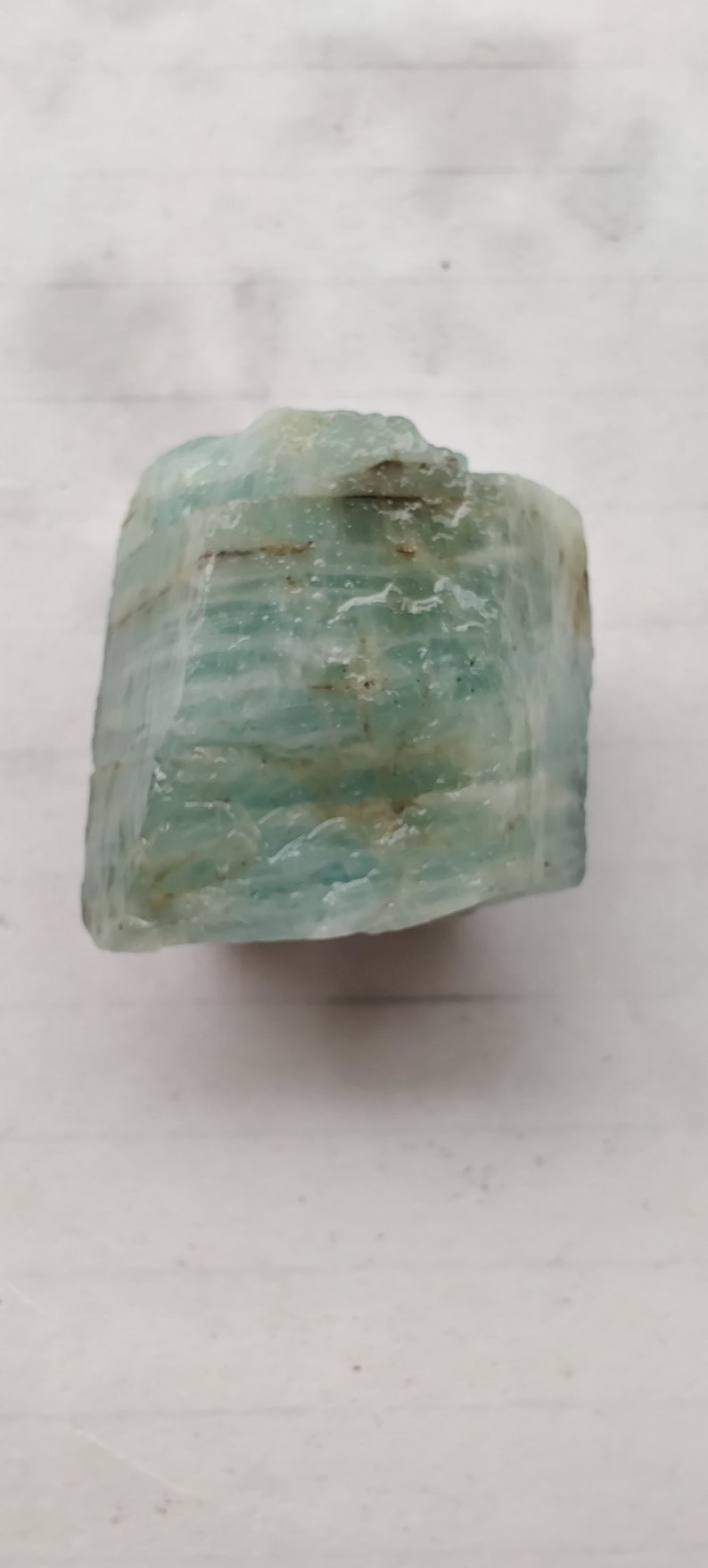 Pode incluir: Um cristal de &aacute;gua-marinha azul claro com uma textura &aacute;spera e natural. O cristal tem uma apar&ecirc;ncia em camadas com estrias vis&iacute;veis.