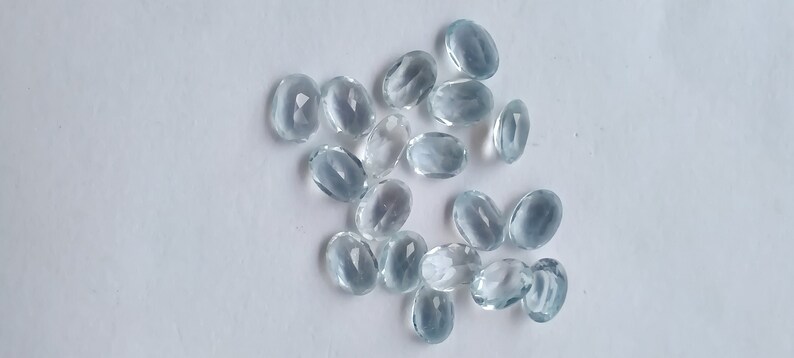 May include: A scattering of oval, pale blue gemstones with a faceted cut, displayed on a white background. The gems vary in size and shape, reflecting light. These gemstones are likely intended for jewellery or craft projects.