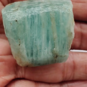 Pode incluir: Um grande cristal de &aacute;gua-marinha bruto de cor verde claro com estrias vis&iacute;veis. O cristal &eacute; transl&uacute;cido e tem uma apar&ecirc;ncia ligeiramente nublada.