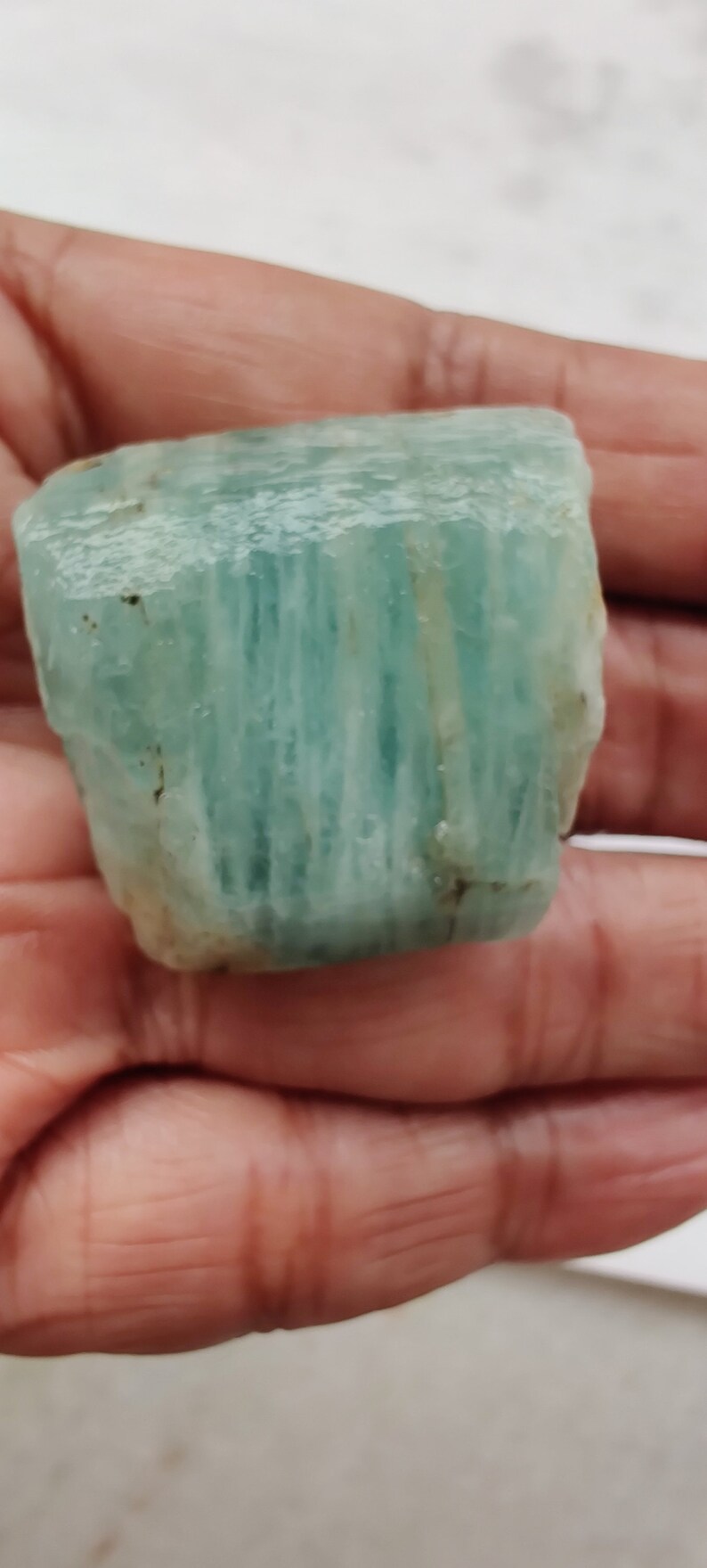 Pode incluir: Um grande cristal de &aacute;gua-marinha bruto de cor verde claro com estrias vis&iacute;veis. O cristal &eacute; transl&uacute;cido e tem uma apar&ecirc;ncia ligeiramente nublada.