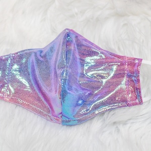Puede incluir: Una mascarilla facial con un acabado brillante e iridiscente en tonos rosa, morado y azul. La mascarilla tiene cintas elásticas blancas para las orejas y está diseñada para cubrir la nariz y la boca. La tela tiene un aspecto texturizado y punteado.
