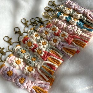 May include: A set of colorful macrame keychains with daisy flower designs and tassels. The keychains are made with a variety of colors, including pink, purple, green, blue, and yellow. Each keychain has a gold-toned metal key ring.