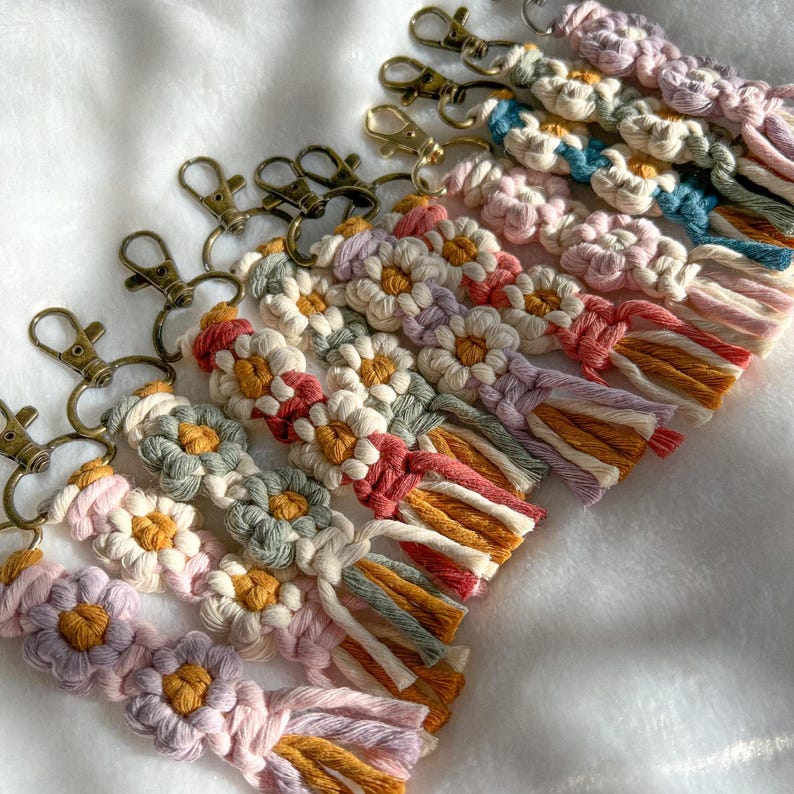 May include: A set of colorful macrame keychains with daisy flower designs and tassels. The keychains are made with a variety of colors, including pink, purple, green, blue, and yellow. Each keychain has a gold-toned metal key ring.