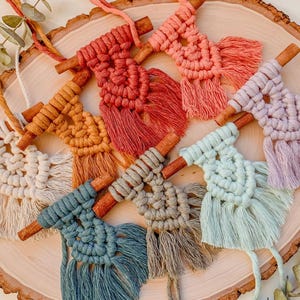 May include: A collection of eight handmade macrame wall hangings with cinnamon sticks. The hangings are in various colours, including white, beige, light blue, teal, grey, light green, orange, and pink. Each hanging has a fringe at the bottom.