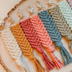 Może przedstawiać: Zestaw siedmiu kolorowych breloków z makramą z srebrnymi kółkami. Breloki wykonane są w różnych kolorach, w tym białym, żółtym, pomarańczowym, czerwonym, niebieskim, zielonym i jasnozielonym. Breloki mają około 25 cm długości.