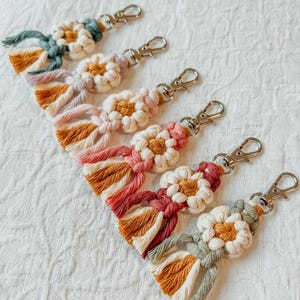 May include: A set of six colorful macrame keychains with daisy-shaped designs. Each keychain features a different color combination of white, pink, red, orange, green, and blue.
