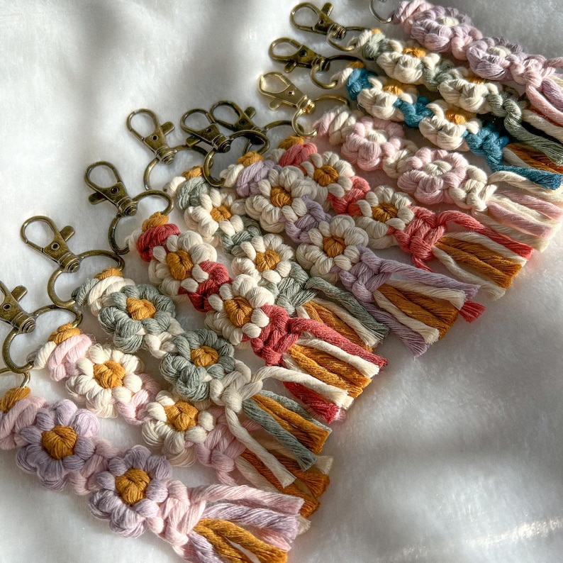 May include: A set of colorful macrame keychains with daisy-like patterns and gold key rings. The keychains are made with a variety of colors, including pink, purple, blue, green, and yellow.