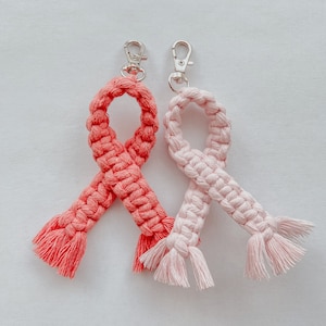 Puede incluir: Dos llaveros de cinta de crochet, uno rosa y otro coral, con anillas de metal plateadas. Las cintas están atadas en forma de lazo.