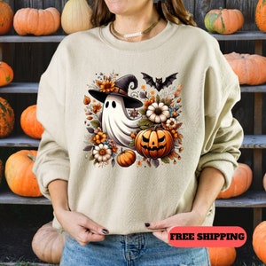 K&ouml;nnte beinhalten: Ein cremefarbener Pullover mit einem Aufdruck eines Geistes, der einen Hexenhut tr&auml;gt und einen K&uuml;rbis h&auml;lt. Der Geist ist von Blumen und Bl&auml;ttern umgeben. Der Aufdruck ist in Orange-, Gelb- und Braunt&ouml;nen gehalten.