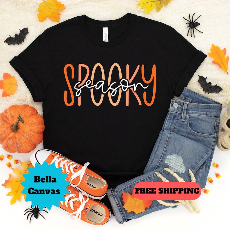 May include: Black t-shirt with the text "Spooky season" in orange and white lettering. The shirt is laid flat on a white surface with fall leaves, a pumpkin, and a pair of orange and white sneakers.