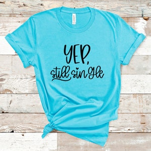 Op de afbeelding: Een turquoise blauw t-shirt met de tekst "Yep, still single" in zwarte letters.