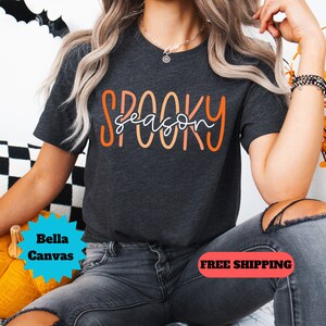 May include: A dark gray t-shirt with the text "Spooky season" in orange and white lettering. The shirt is being worn by a person with long blonde hair and is sitting on a yellow pumpkin.