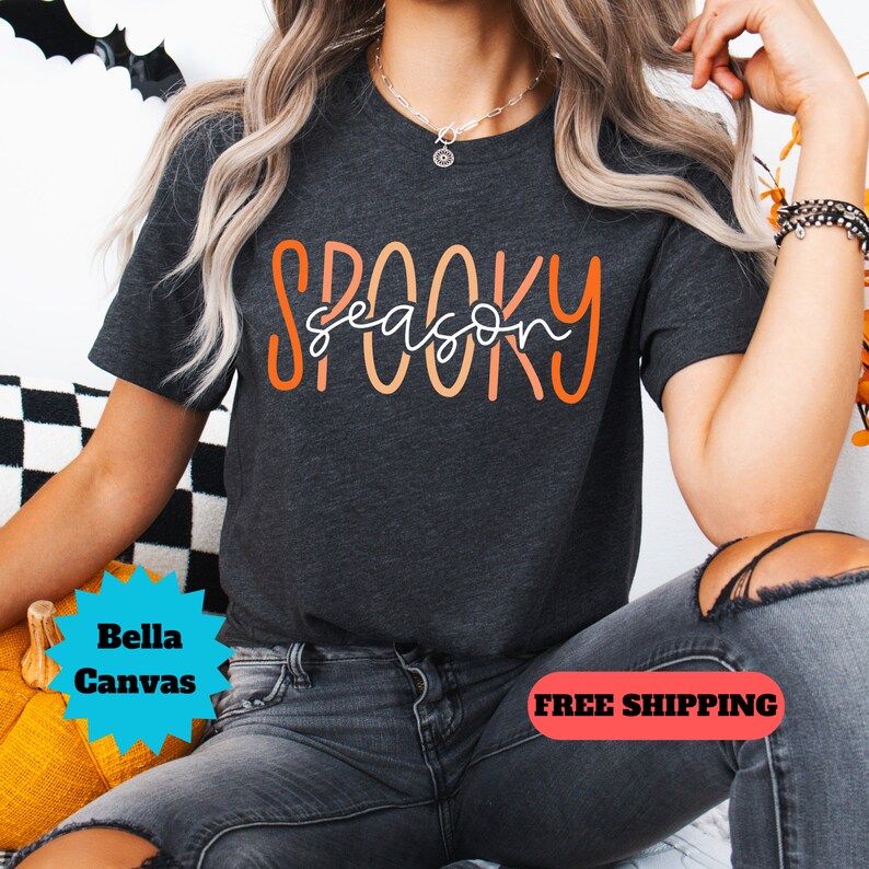 May include: A dark gray t-shirt with the text "Spooky season" in orange and white lettering. The shirt is being worn by a person with long blonde hair and is sitting on a yellow pumpkin.