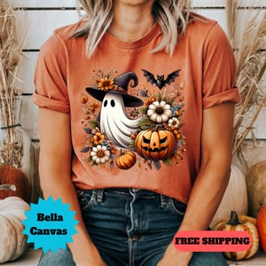 K&ouml;nnte beinhalten: Ein orangefarbenes T-Shirt mit einem wei&szlig;en Geist, der einen Hexenhut tr&auml;gt und einen K&uuml;rbis h&auml;lt. Der Geist ist von Blumen und einer Fledermaus umgeben. Das Shirt hat den Text "Bella Canvas" unten links.