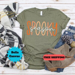 May include: A green t-shirt with the text "Spooky Season" in orange and pink lettering. The shirt is laying on a wooden surface with a pair of brown ankle boots and a blanket.