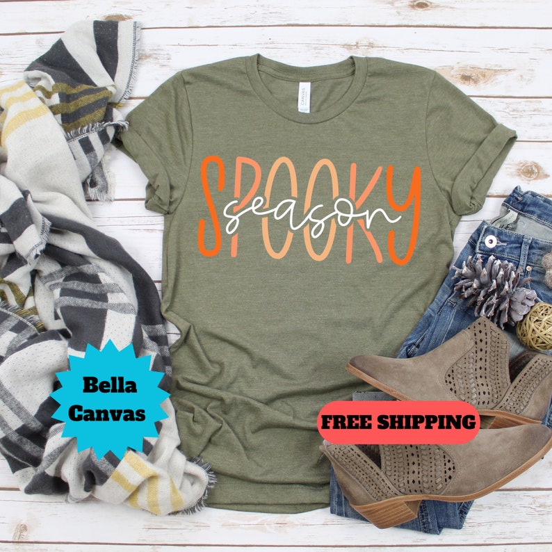 May include: A green t-shirt with the text "Spooky Season" in orange and pink lettering. The shirt is laying on a wooden surface with a pair of brown ankle boots and a blanket.
