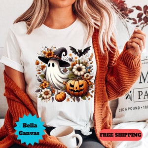 K&ouml;nnte beinhalten: Wei&szlig;es T-Shirt mit einem Aufdruck eines Geistes, der einen Hexenhut tr&auml;gt und einen K&uuml;rbis h&auml;lt. Der Geist ist von Blumen und Bl&auml;ttern umgeben. Der Aufdruck ist im Cartoon-Stil.