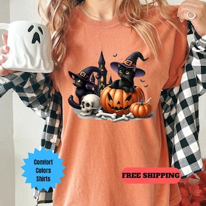 May include: Orange t-shirt with a black cat wearing a witch hat sitting inside a jack-o-lantern. The cat is holding a skull and bones. The text "FREE SHIPPING" is at the bottom of the image.