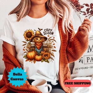May include: White t-shirt featuring a fall-themed graphic with a scarecrow, pumpkins, and sunflowers. The text "COZY SEASON" is above the scarecrow. The shirt is a Bella Canvas brand.