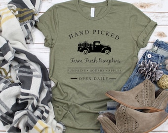 May include: A green t-shirt with the text "Hand Picked Farm Fresh Pumpkins" and a black silhouette of a truck with pumpkins in the bed. The text below the truck reads "Pumpkins Gourds Apples - Open Daily - "
