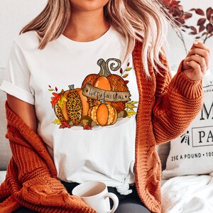 May include: White t-shirt with a graphic of three pumpkins. The pumpkins are orange and brown with a leopard print design. The text "Happy Fall Y'all" is on a banner in front of the pumpkins.