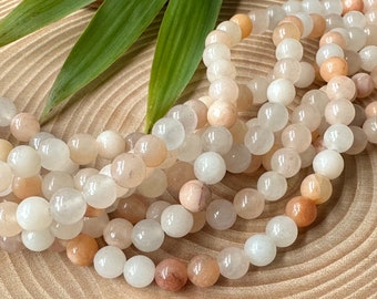May include: A strand of smooth, round beads in varying shades of translucent white, cream, and soft peach. The beads are polished and reflective, arranged in a loose, coiled formation, showcasing a natural variation in color and opacity throughout.