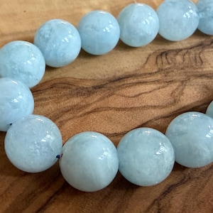 Könnte beinhalten: Eine Kette aus runden, polierten hellblauen Aquamarinperlen. Jede Perle hat eine glatte, glänzende Oberfläche mit subtilen, wolkenartigen Einschlüssen im Inneren und natürlichen Farbvariationen, angeordnet in einer geschwungenen Linie.