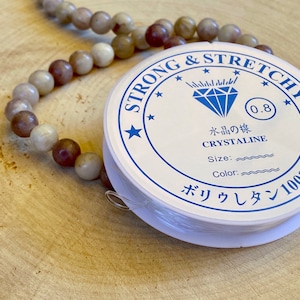 May include: A white spool of strong and stretchy elastic cord for jewelry making. The label reads "Strong & Stretchy", "0.8", "Crystaline", "Size:", "Color:", and "ボリウレタン100%".