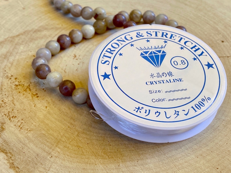 May include: A white spool of strong and stretchy elastic cord for jewelry making. The label reads "Strong & Stretchy", "0.8", "Crystaline", "Size:", "Color:", and "ボリウレタン100%".