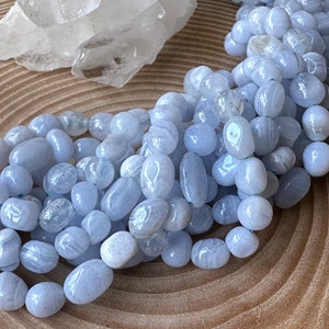 Könnte beinhalten: Eine Ansammlung von glatten, getrommelten hellblauen Spitzenachat-Perlen mit sichtbaren weißen Bändern und durchscheinenden Bereichen. Die Perlen sind oval und abgerundet geformt und weisen eine polierte Oberfläche sowie eine kühle, hellblaue Farbpalette auf.
