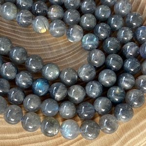 May include: A collection of round, polished labradorite beads in shades of dark gray and blue. Each bead displays a smooth, reflective surface with subtle internal flashes of iridescent light, arranged in a loose, overlapping circular pattern.