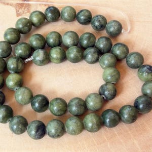 May include: A strand of round, smooth beads in varying shades of forest green and olive. Each bead features a polished surface with natural dark inclusions and subtle marbling patterns, strung together on a clear, flexible filament cord.