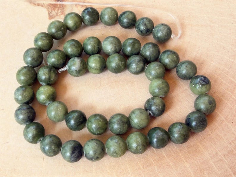 May include: A strand of round, smooth beads in varying shades of forest green and olive. Each bead features a polished surface with natural dark inclusions and subtle marbling patterns, strung together on a clear, flexible filament cord.