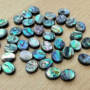May include: A collection of oval-shaped abalone shell beads with iridescent green, blue, and purple hues. The beads have a smooth, polished surface and are strung on a black cord.