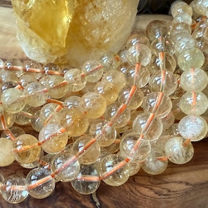 May include: A strand of translucent, pale yellow citrine beads arranged in multiple loops. The spherical beads are strung on a thin, bright orange cord, showcasing internal natural inclusions and a polished, reflective surface texture.
