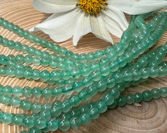 May include: A strand of smooth, round, translucent light green aventurine beads. The beads are uniform in size and shape, featuring a soft, polished finish that catches the light. The strand is arranged in multiple loops, showcasing the consistent pale green color and subtle internal variations of the natural stone material.