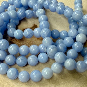 Könnte beinhalten: Stränge hellblauer, runder Perlen mit marmoriertem Aussehen. Die Perlen sind aufgefädelt und bilden eine Halskette oder ein Armband. Die Perlen haben eine weiche, pastellblaue Farbe.