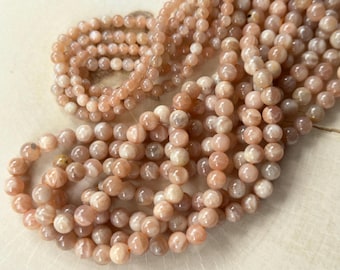 May include: A collection of round, polished peach-colored moonstone beads arranged in loose strands. Each bead features a smooth, glossy surface with subtle internal light reflections and variations in tone ranging from pale cream to soft orange.