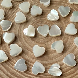 Op de afbeelding: Een verzameling witte parelmoer hartvormige kralen. De kralen zijn in een cirkelvormige opstelling op een houten oppervlak.